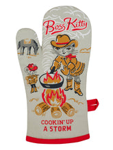 Load image into Gallery viewer, Grey/blue oven mitt with an illustration of a cat wearing a cowgirl outfit cooking on a bonfire. in the background her horse is grazing and another kitty is collecting wood for the fire. The text “Boss Kitty” s at the top, and “Cookin’ Up A Storm” is at the bottom.