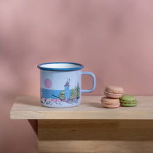 Load image into Gallery viewer, Enamel mug on a wooden shelf The mug has a pale blue background with a scenic design of moomin family with their house and the sea and a sunset.