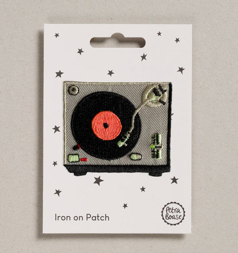 The iron on patch is pictured on its backing card with little stars on.  The patch depicts a turntable from above, with a record playing.