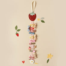Load image into Gallery viewer, This hair clip hanger has a ribbon hanging hook at the top, below which is a glittery fabric strawberry.  Attached is a length of soft white ribbon with little red dots which is the perfect size for clipping on hair clips for neat and accessible storage.  At the bottom of the ribbon length there is a little felt daisy.  The clip hanger is shown here with 12 different hair clips on it.
