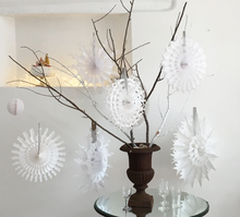 Load image into Gallery viewer, White paper snowflake decorations displayed on a glass table with a dark brown urn vase and bare branches against a white wall.