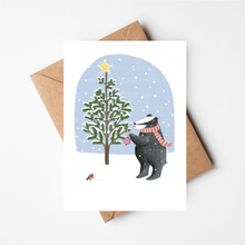 Load image into Gallery viewer, A white greeting card with a winter illustration of a badger in a red-and-white striped scarf decorating a Christmas tree, shown with a brown kraft envelope on a light gray background.