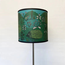 Load image into Gallery viewer, a lamp fitting lamshade in green , blue, black and white featuring 5 hedgehogs seen in a garden with flowers.  There is a black trim to the top and the bottom of the shade.  The shade is pictured on a modern looking metal lamp stand

