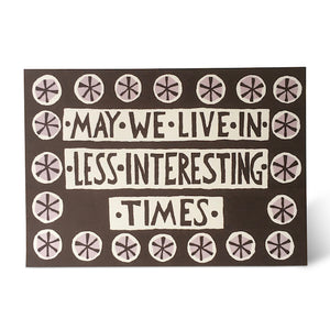 Greetings card with dark grey backround and white, hand draen lettering which reads "may we live in less interesting times".  Arounf the edges of the card is a simple border design of sehmented circles.