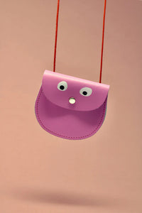 bright pink leather mini bag, with a red cord carrying string.  Simple design with stitching around the curved  sides. The bag has a front flap with a small silver popper, which also looks like a nose as there are 2 simple white and black googly eyes painted on to the leather, making the bag has a little face.