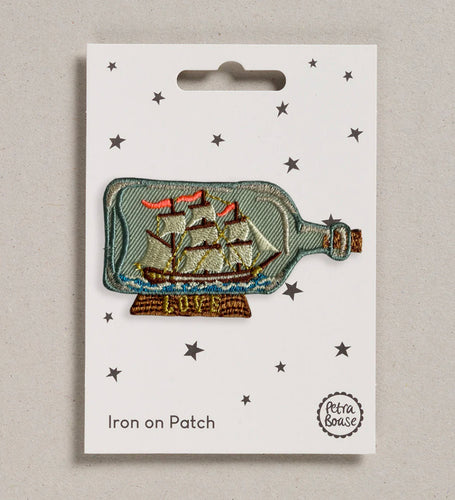 The iron on patch of a bittle is shown on its backing card.  The ship has three masts with sails and is mounted on a little stand with the words “LOVE” on it.