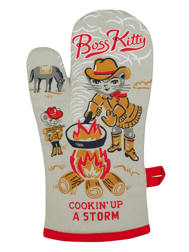 Grey/blue oven mitt with an illustration of a cat wearing a cowgirl outfit cooking on a bonfire. in the background her horse is grazing and another kitty is collecting wood for the fire. The text “Boss Kitty” s at the top, and “Cookin’ Up A Storm” is at the bottom.