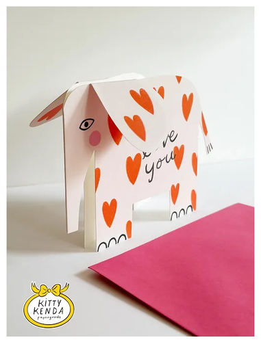 cut out shaped card of  an elephant with sticking out 3D ears.  \The elephant is white with red hearts all over and cute pink cheeks