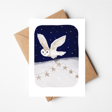Load image into Gallery viewer, A white greeting card with a rounded rectangular illustration of a white barn owl flying across a snowy night scene under a dark blue sky, shown with a brown kraft envelope on a light gray background.
