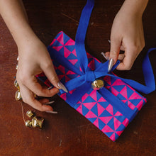 Load image into Gallery viewer, A close-up of hands wrapping a gift box with bright blue and pink geometric patterned paper and a royal blue ribbon tied in a bow with small gold bells, set on a dark wooden table.
