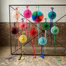 Load image into Gallery viewer, A set of colorful paper fan decorations hanging from a black wire grid frame in an industrial room with a brick wall and concrete floor.
