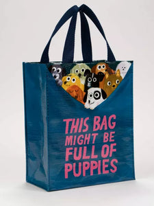Cute little tote bag by blue Q featuring an illustration by John Bond. The bag is mostly dark petrol blue with bright pink lettering which reads “THIS BAG MIGHT BE FULL OF PUPPIES”. At the top edge of the bag 9 little assorted puppies can be seen, as if they are pulling the rim of the bag down and are inside.