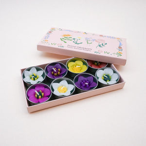 Eight flower shaped tea lights can be seen in a pretty pink box with a floral design   And the words”hana blossom.  Flowers Scented Tealights”.  Each candle is delicately made to look like a different flower, in pinks, yellows, white, purple and red.