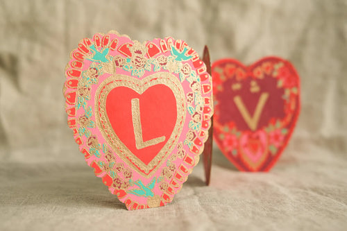 The beautiful red, pink, turquoise and gold, hand printed, heart shape concertina card can be seen open, with the letter L on the first side and the fold with the V visible on the background.  The card has 4 hearts i. A row spelling LOVE.