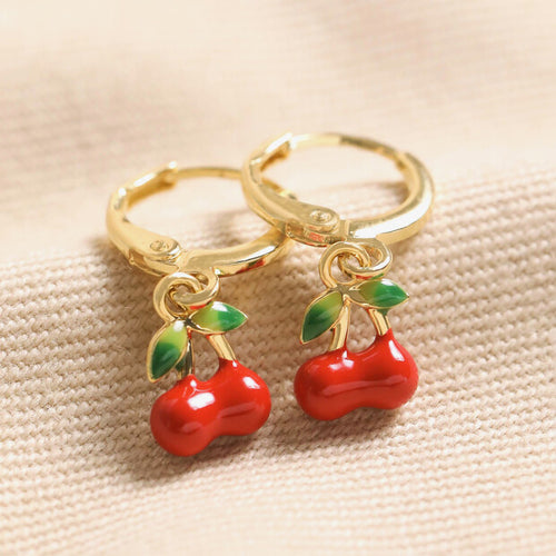 Gold huggie hoop earrings with small enamel red cherry charms hanging from them.  The cherries also have a little pair of green leaves on each of them.