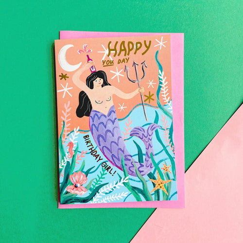 Card featuring a mermaid holding a bottle with liquid spurting out on top of her head.  She has black hair and a purple fishy tale.  The words HAPPY YOU DAY, are in gold at the top of the card, and also the words BIRTHDAY GIRL are seen beside her tail.