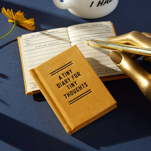 the tiny mustard coloured diary can be seen with its cover with the words in black capitals saying 'A TINY DIARY FOR THOUGHTS'.  Underneathe the closed diary is an open one revelaing the inside sheets which are lined with a place for the date and to number the topic and 'brilliant realization'