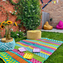 Load image into Gallery viewer, the bright woven plastic rug is shown on a lawn with poufs on it.
