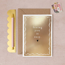 Load image into Gallery viewer, In the centre of a shiny gold card with decorative curly boarder, the words &#39;loving you is easy&#39; in a simple white font, finihsed with a small black heart illustration. 
