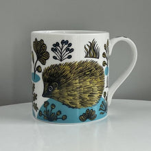 Load image into Gallery viewer, Two beautifully illustrated hedgehogs surrounded by plants and flowers. A white mug with mustard yellow and blue designs
