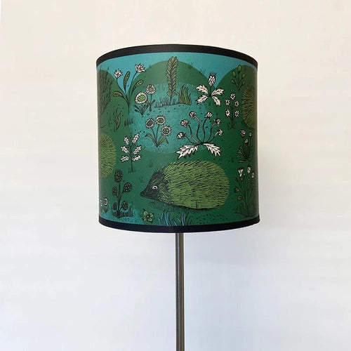 a lamp fitting lamshade in green , blue, black and white featuring 5 hedgehogs seen in a garden with flowers.  There is a black trim to the top and the bottom of the shade.  The shade is pictured on a modern looking metal lamp stand