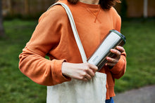 Load image into Gallery viewer, a person can be seen placing the travel tumbler in a cotton tote bag on their shoulder
