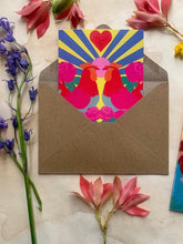 Load image into Gallery viewer, the card is seen coming out of the kraft envelope.  There are two roosters facing each othe and behind them is a sunrise with a large heart in the middle of the rays, and they are surrounded by roses.  The card is extremely brightly coloured with green, blue red , pink  and yellow
