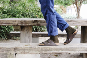 A cropped image of a persons lewer legs and feet, walking along a wooden bench wearing blue trousers, khaki socks and sandles. 