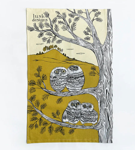 this tea towel is printed in green and black and white.  It features 4 baby owls sitting in pairs in 2 branches of a tree.  A green hill with trees on top can be seen on the background.