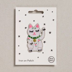 A sweet little waving lucky cat, white with pink ears and a green collar with a gold pendant on it.  There is also three little pink and green flowers on the front of the cat.  The patch is shown on its backing card which has small stars on it.